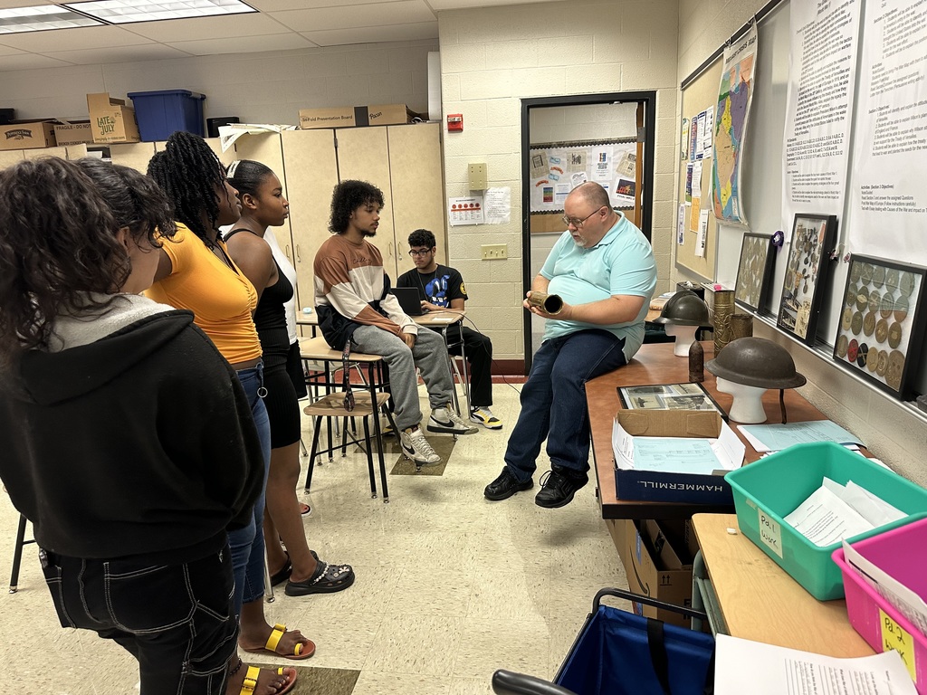 A group of students attentively listens to an instructor demonstrating historical artifacts in a classroom setting.