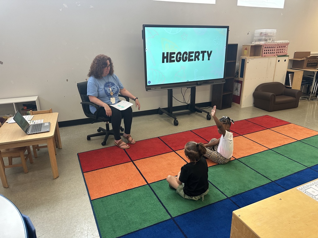 A teacher teaching two young students in a classroom. One student is raising her hand.