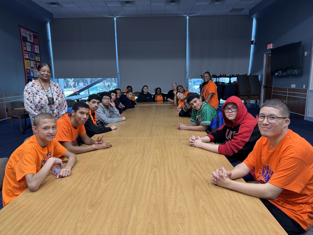 Group of students and one adult sitting at a large table. Two other adults are standing next to the students.