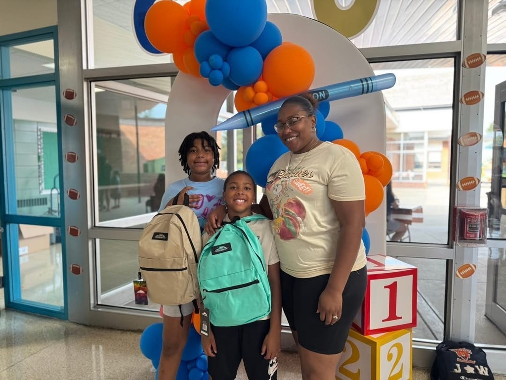 An adult posing with two students in front of a backdrop at Goode PreK-8's Back to School Night. The two students are holding up bookbags.