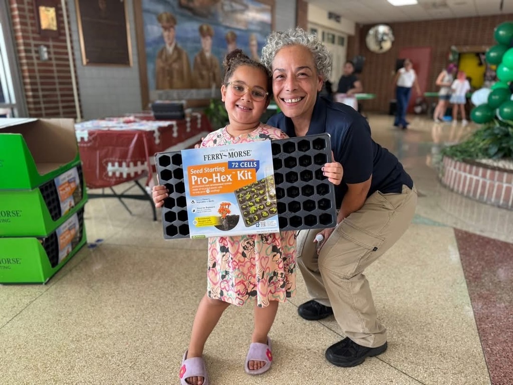 A district staff member bending down next to a student holding up a book titled "Seed Starting Pro-Hex Kit."