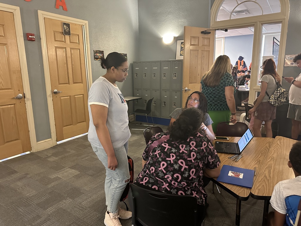 An adult is standing up looking at a laptop screen while a district staff member is talking to her. Another adult is sitting at the table. 