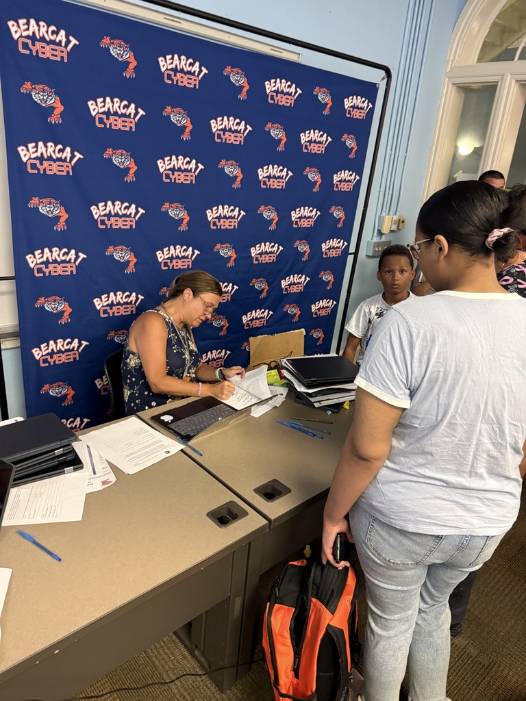 A district staff member writing on a piece of paper with an adult standing in front of the table. A child is in the background.