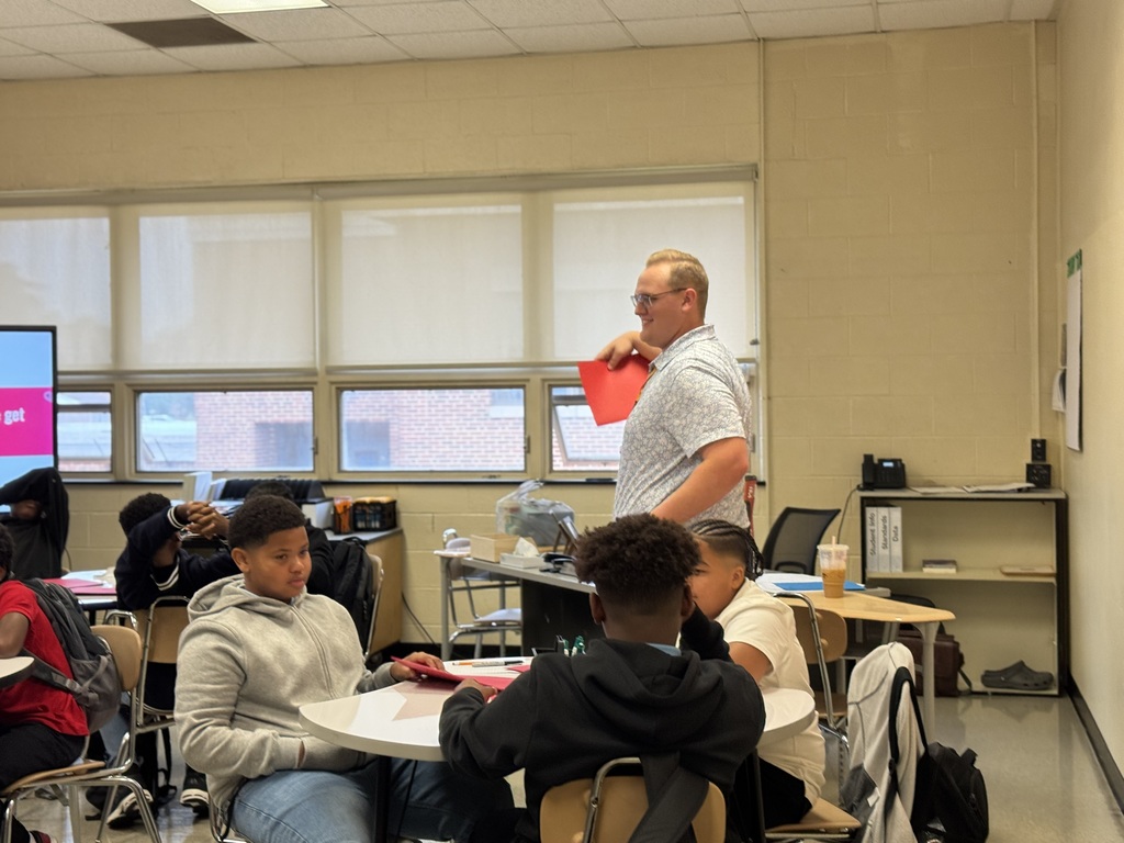 A teacher holding up a piece of paper in a classroom with students sitting at tables.