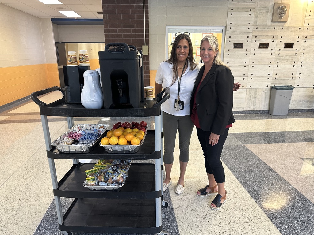 Two district staff members pushing a cart with fruit, coffee, and other snacks on it.