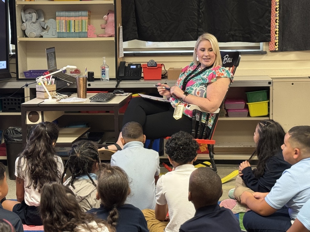 A teacher sitting in a chair with students sitting on the carpet looking up at her.