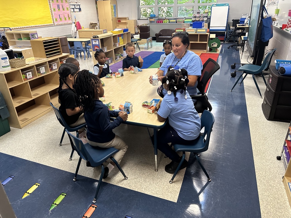 A teacher and 7 students sitting at a table. The teacher is pointing to a milk carton.