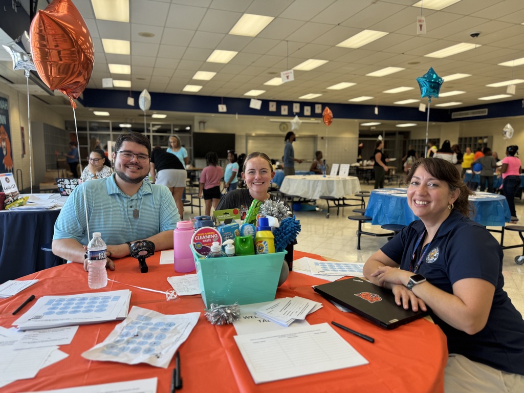 Three district staff members sitting at an orange table with an orange balloon, multiple papers and a basket of cleaning supplies on it. People can be seen in the background.