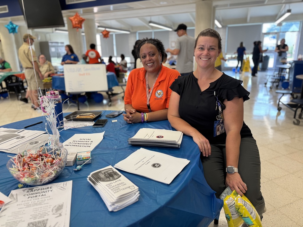 Two district staff members sitting at a table with multiple papers, brochures and candy on it during Hannah Penn's recent Back to School Night. People can be seen in the background.