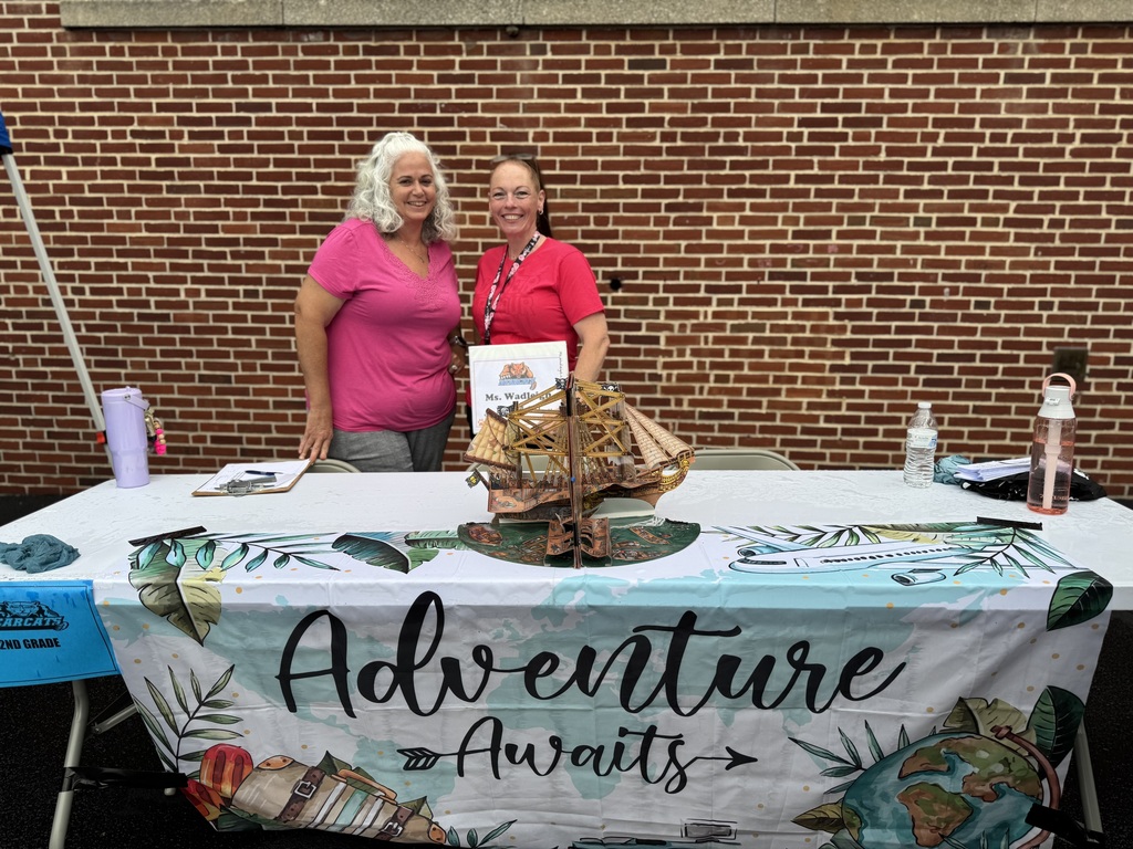 Two district staff members posing behind a table that says "Adventure Awaits" while standing in front of a brick wall.