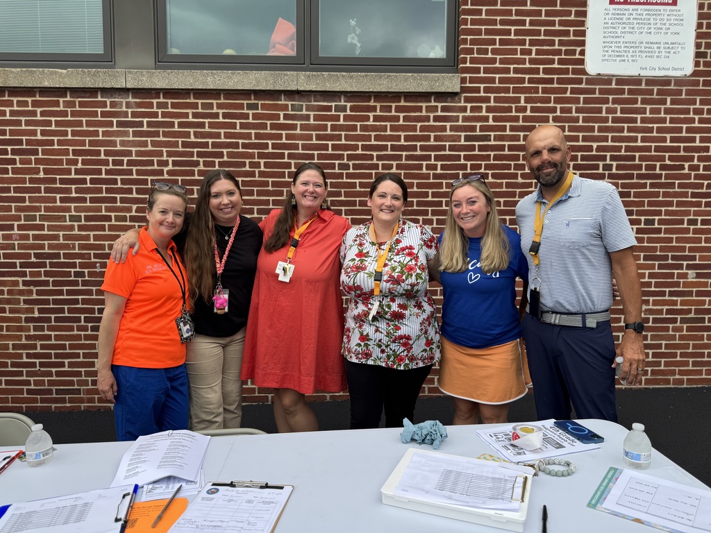 Six district staff members posing behind a table with papers and pens on it while standing in front of a brick wall.