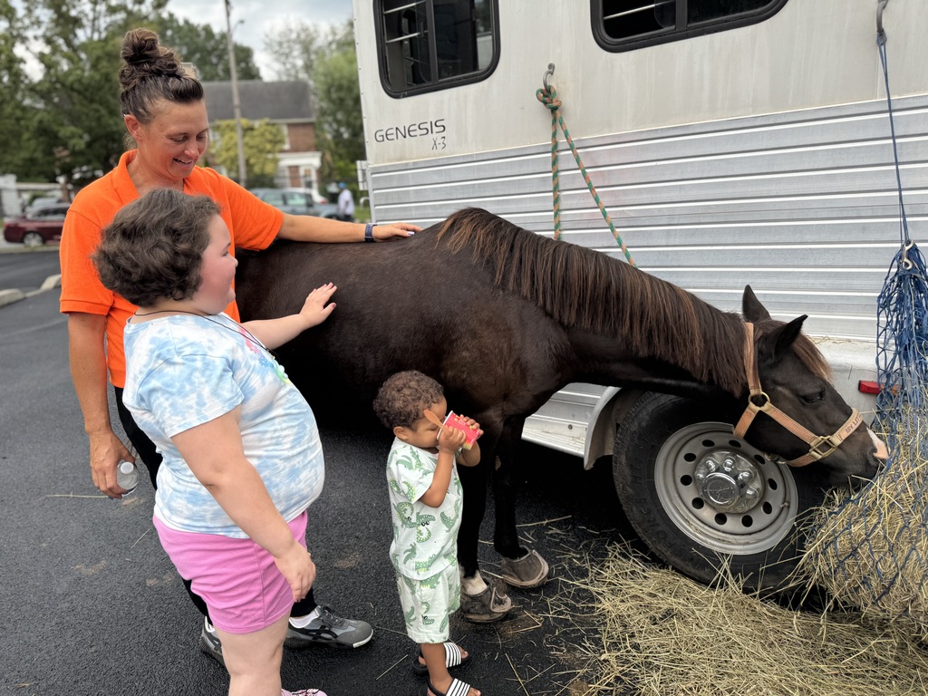 An adult and a student are petting a horse while a small child is drinking out of a cup that has a spoon in it.