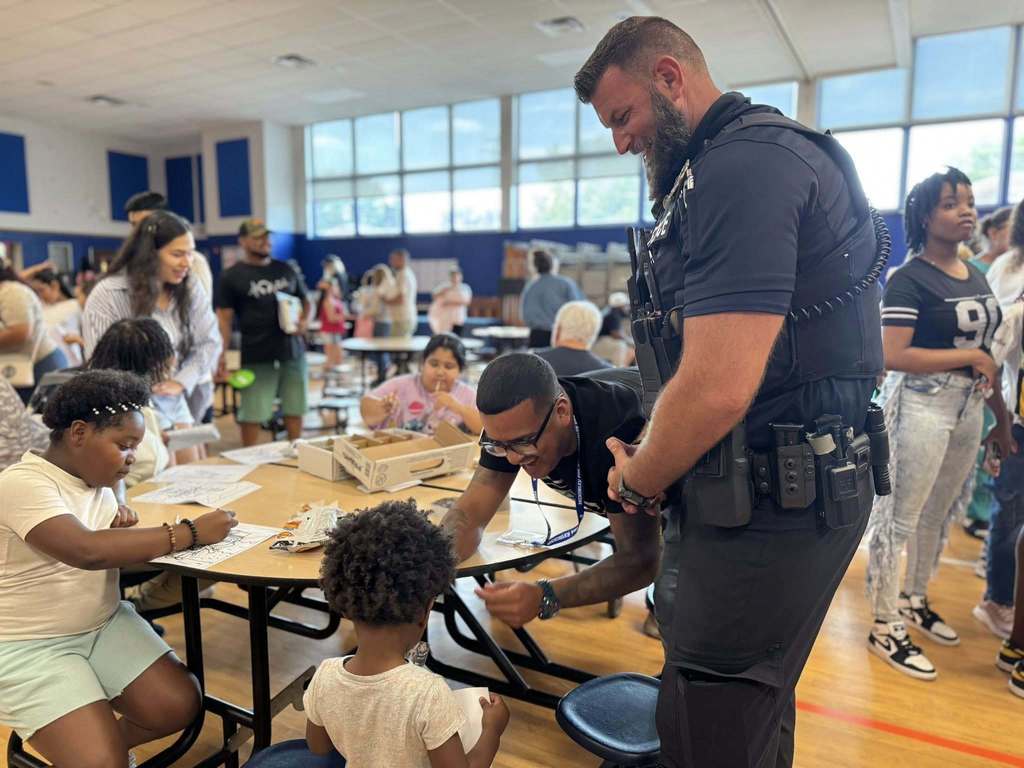 District staff looking down at a student who is standing near a table and smiling. People can be seen in the background.