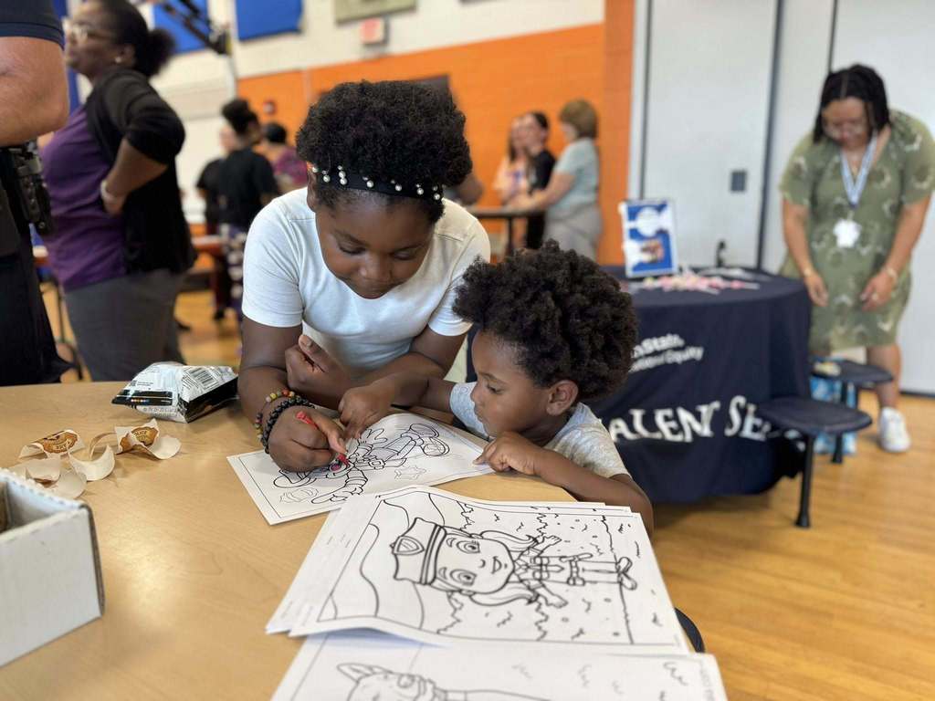 Two children coloring with a crayon on a table. People can be seen in the background. 
