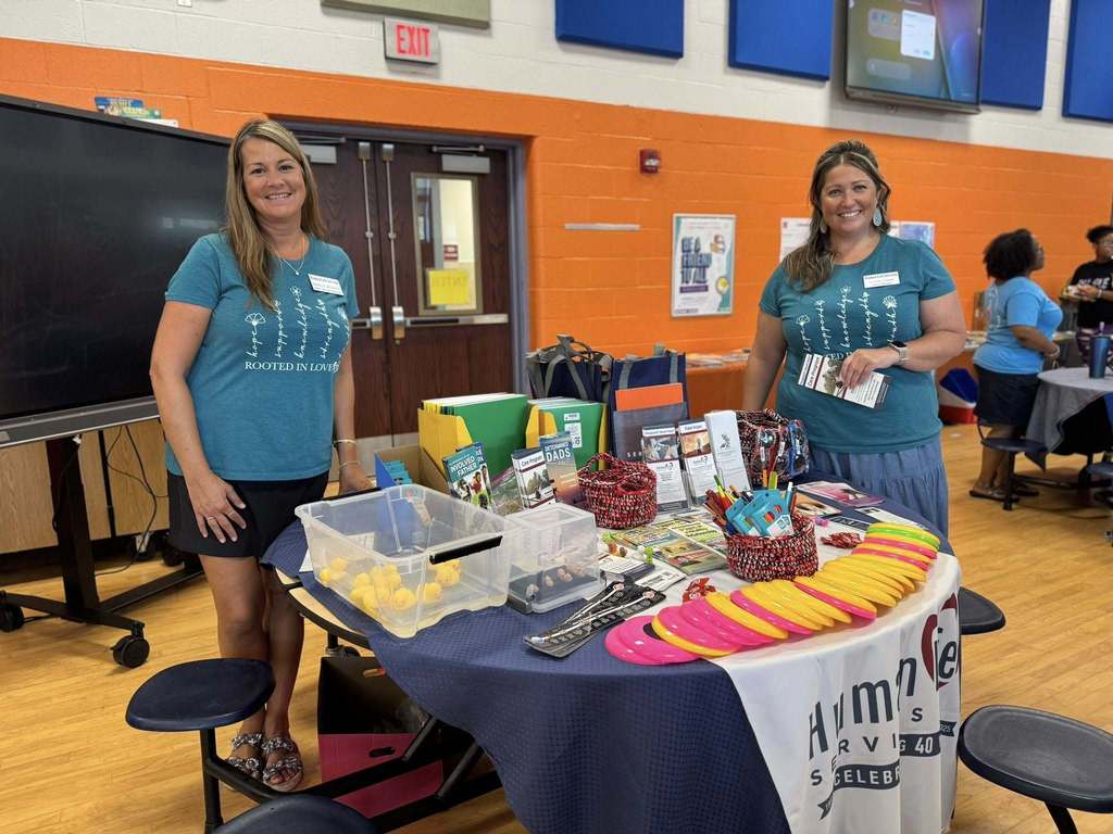 Two adults posing by a table that has multiple items on it. Some people can be seen in the background.