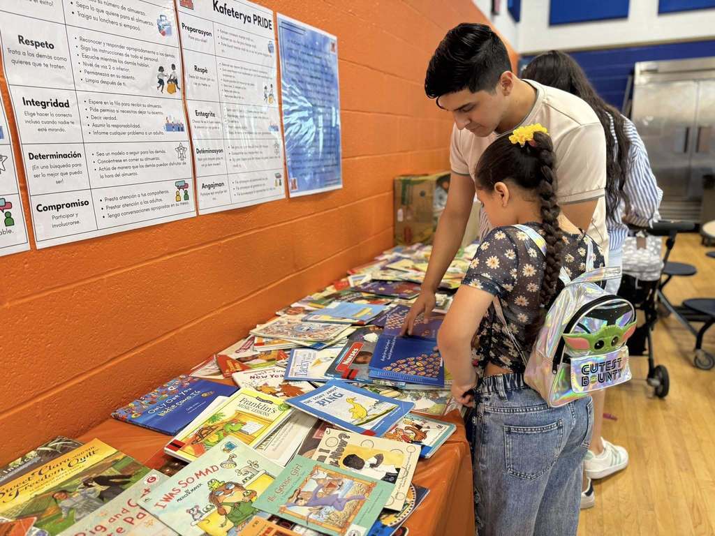 An adult and a young child looking at a book at a table that is filled with books. A person can be seen in the background.