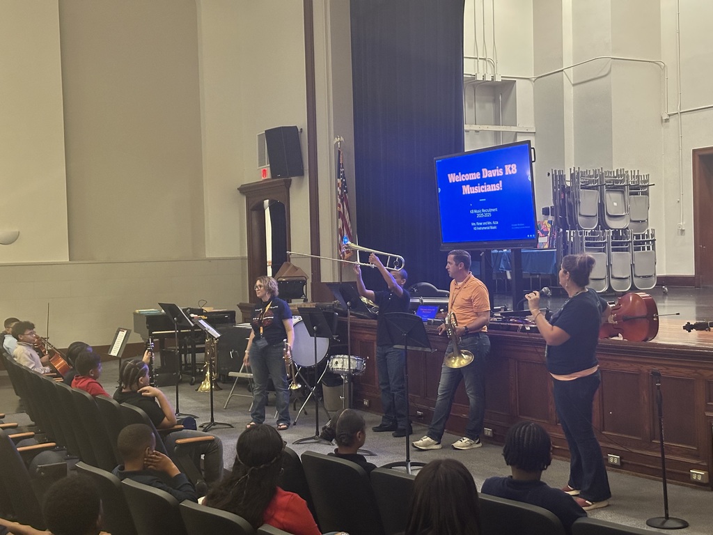 A student holding up an instrument for the crowd to see during the district's annual K-8 Music Recruitment Assembly. One of the district staff members is speaking into a microphone.