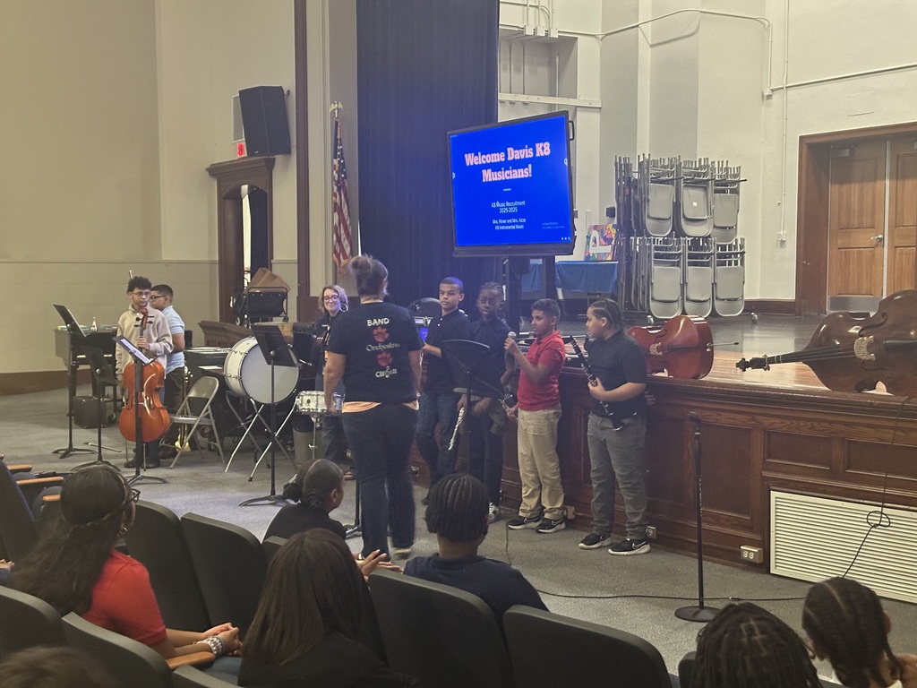 A student holding a microphone while standing in a line with other students during the district's annual K-8 Music Recruitment Assembly.