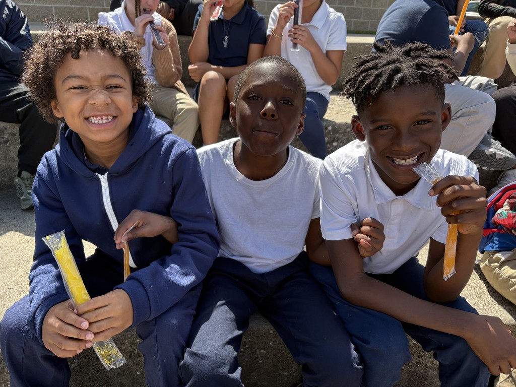 Three students holding popsicles in their hands and smiling. Other students can be seen in the background.