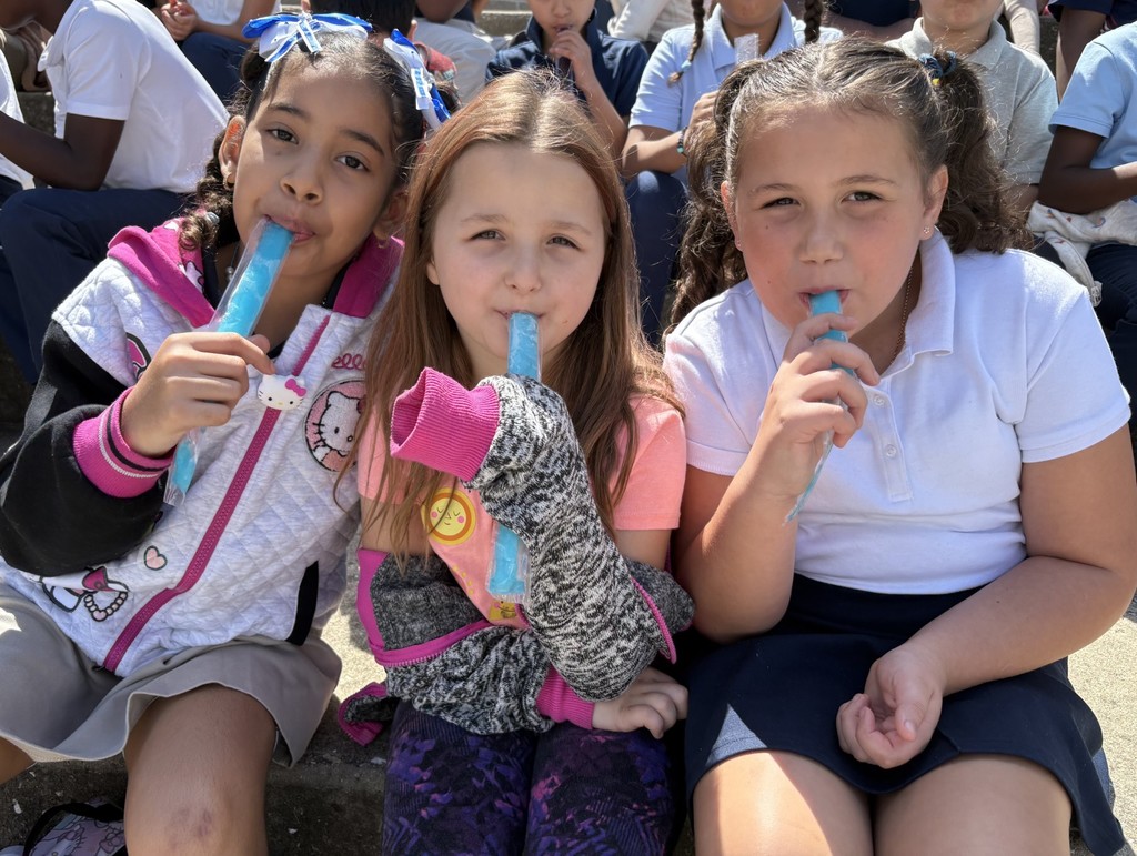 Three students holding popsicles in their hands and smiling. Other students can be seen in the background.