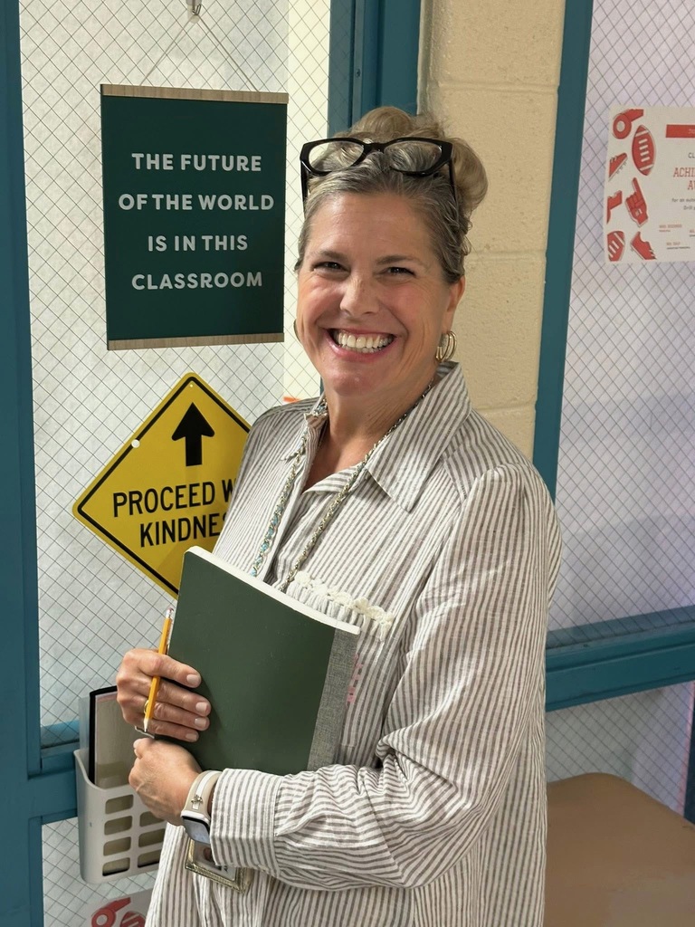 A district staff member smiling for a photo while standing in front of a sign that says "The Future of the world is in this classroom". There is another sign that says "Proceed with Kindness". 