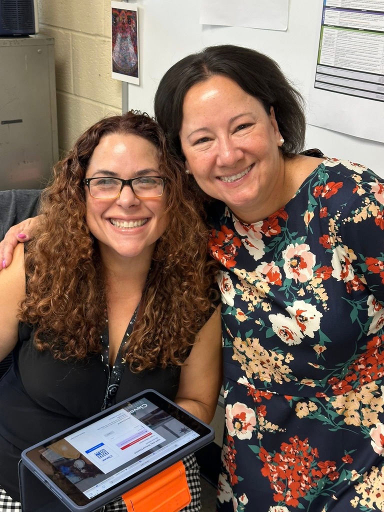 District staff members posing together for a photo. One of the district staff members is holding an iPad. The other district staff member has her hand around the district staff member that is holding the iPad. 