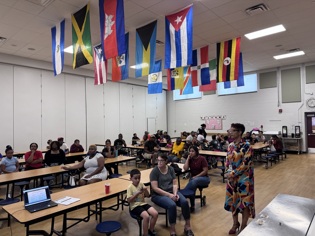 Jackson PreK-8 Principal and a group of people sitting down in a school cafeteria. Multiple flags are hanging from the ceiling.