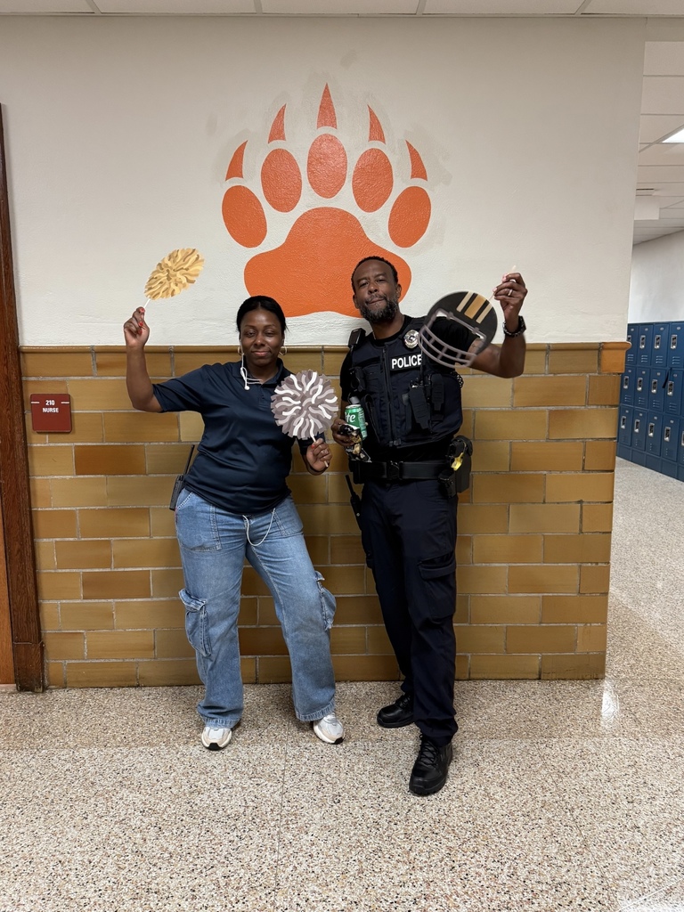 A district staff member in casual attire and a school police officer posing together in a school hallway, each holding decorative items in front of a bear paw mural.