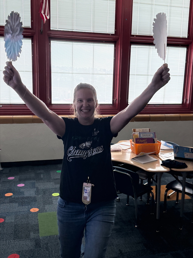 A district staff member standing in a classroom, joyfully holding up two colorful paper fans. The background shows desks and educational materials.