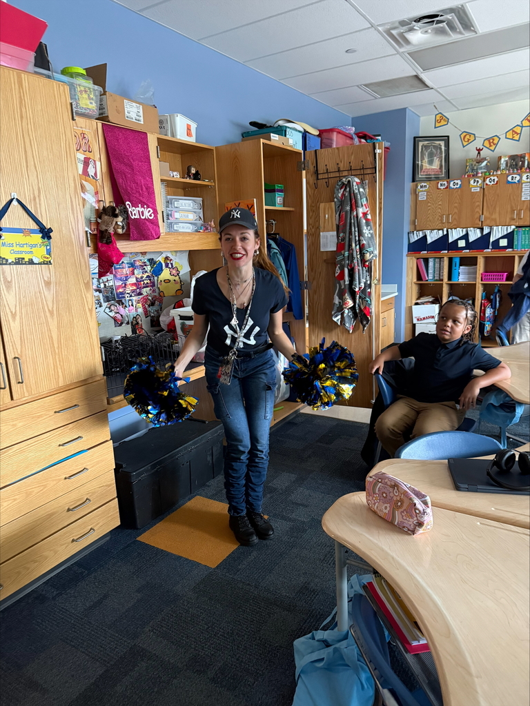 A district staff member is holding blue and gold pom-poms in both of her hands, while standing inside of a classroom. A student is sitting at a desk in the background, looking at the district staff member.