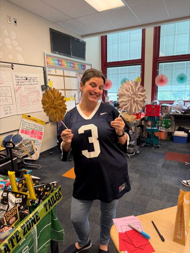 A district staff member is holding two paper pom-poms in her hands while standing in a classroom decorated with colorful hanging paper fans. A table with snacks and football-themed decorations is nearby.
