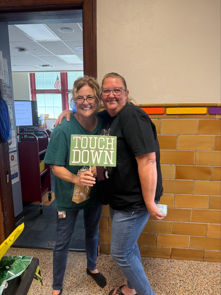 Davis PreK-8 Principal, Ms. Sweitzer and a district staff member are posing joyfully in a school hallway, one holding a green "TOUCH DOWN" sign. Bright classroom decor and a flag are in the background.
