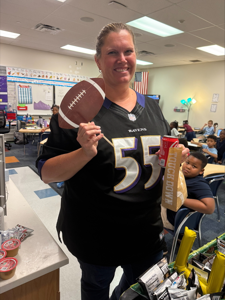 A district staff member in a Ravens jersey holding a football and snacks in a classroom decorated for a football-themed event.