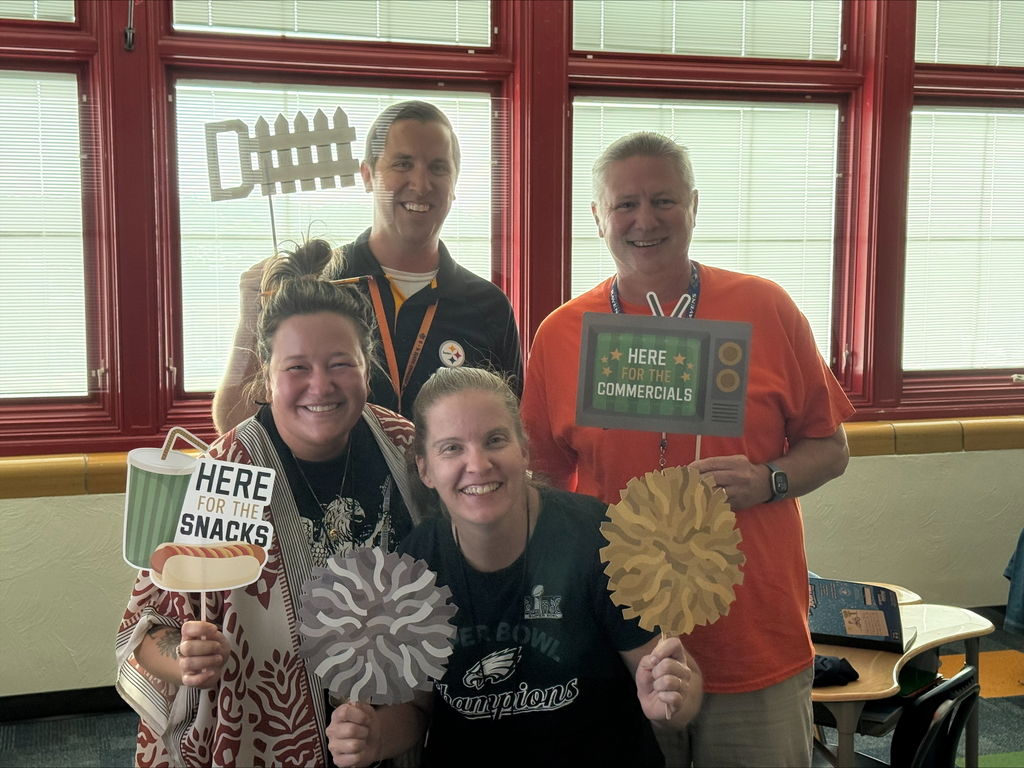 A group of district staff members posing with playful signs, including "Here for Snacks" and "Here for the Commercials," in a bright room.
