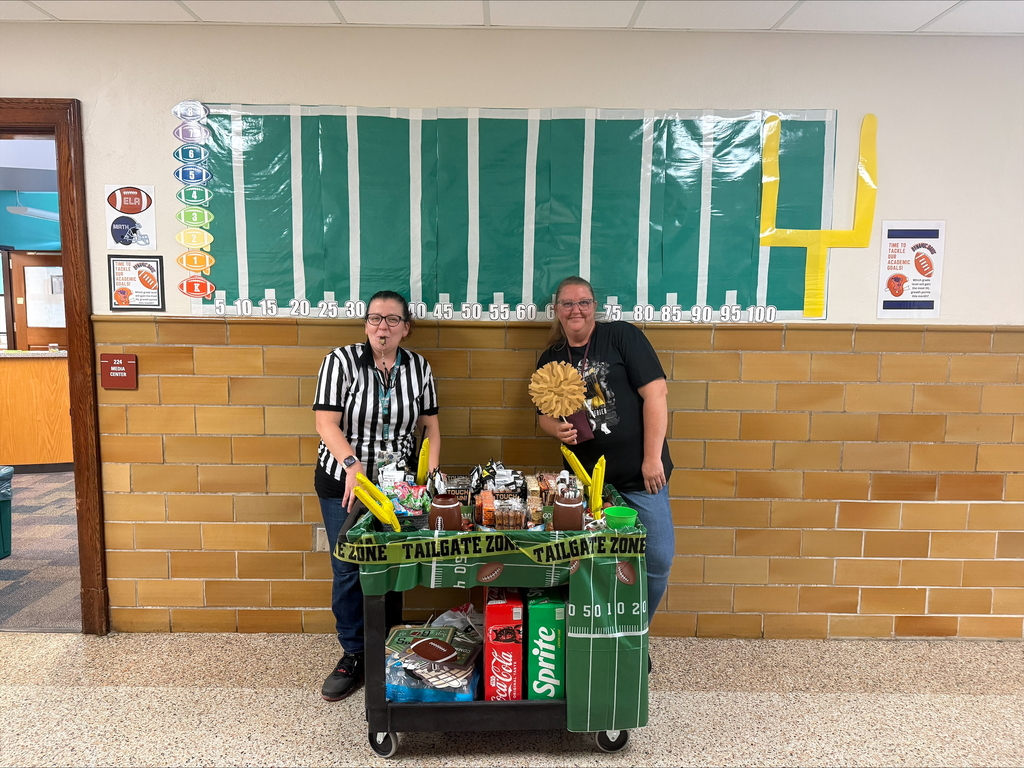 Davis PreK-8 Principal, Ms. Sweitzer and a district staff member are posing in a school hallway with a football-themed snack cart labeled 'Tailgate Zone,' filled with snacks and drinks. One is dressed as a referee, and the other holds a pom-pom. Behind them, a bulletin board is decorated like a football field with yard markers and a yellow goalpost.
