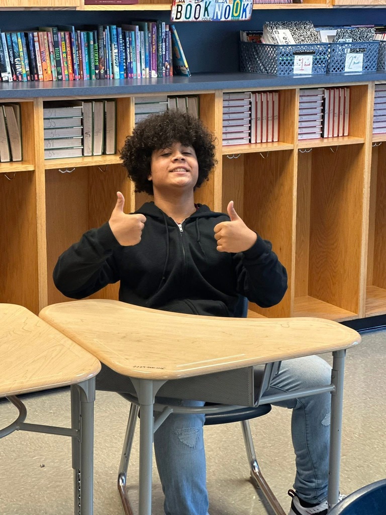 A student is giving a thumbs up while sitting at a triangular desk in a classroom with bookshelves in the background.