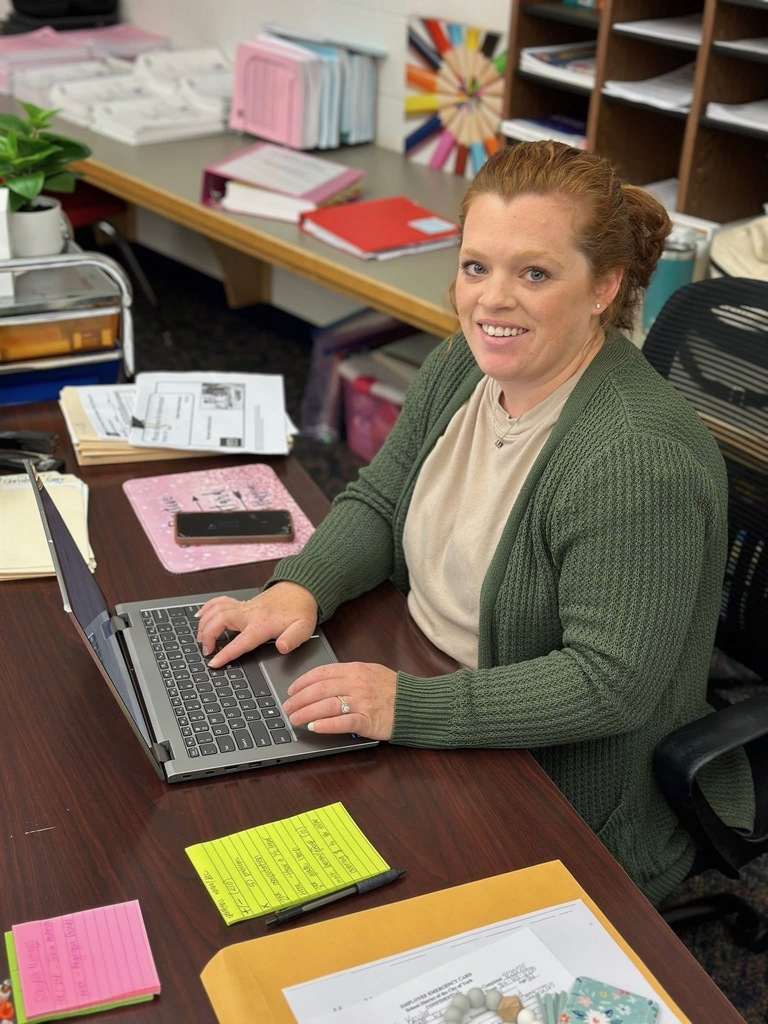 A district staff member is typing on a laptop, surrounded by colorful notepads and office supplies on a wooden desk.