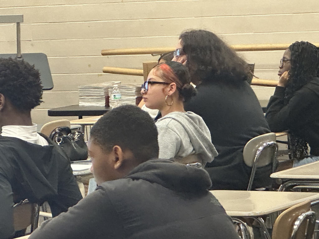 A classroom scene with students seated at desks, focused on a lesson, with textbooks and a music stand in the background.