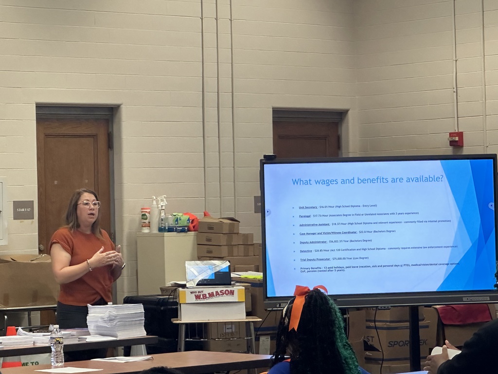 A woman presents information on wages and benefits in a classroom, with a large screen displaying job details and requirements.