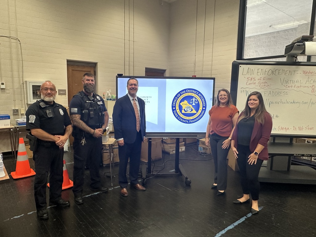 A group of five people stands indoors, smiling. Two are in police uniforms, one in a suit, and two women in casual office attire. A digital screen displays a logo. The setting appears professional, with large whiteboards and traffic cones in the background, suggesting a meeting or presentation.