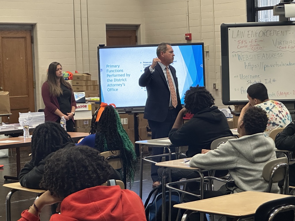 A classroom setting with a speaker presenting to students, while a presentation on the screen outlines district attorney functions.