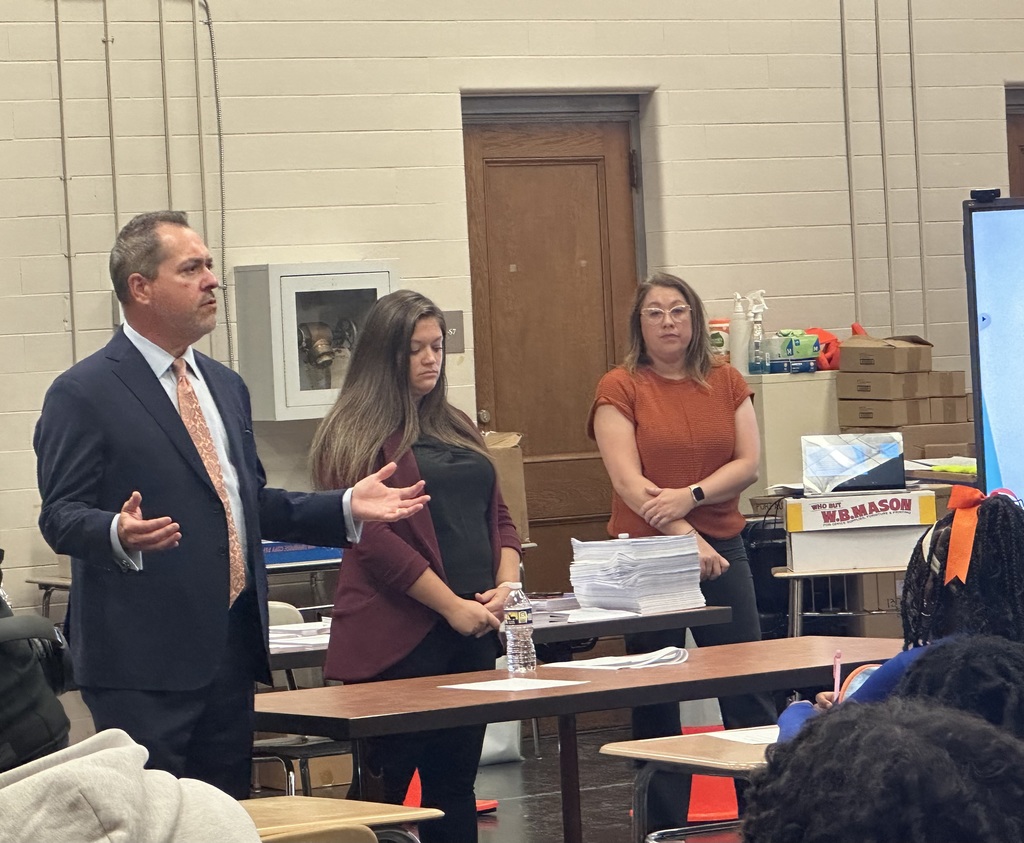 A speaker in a suit gestures while presenting to two women in a classroom filled with desks and educational materials.