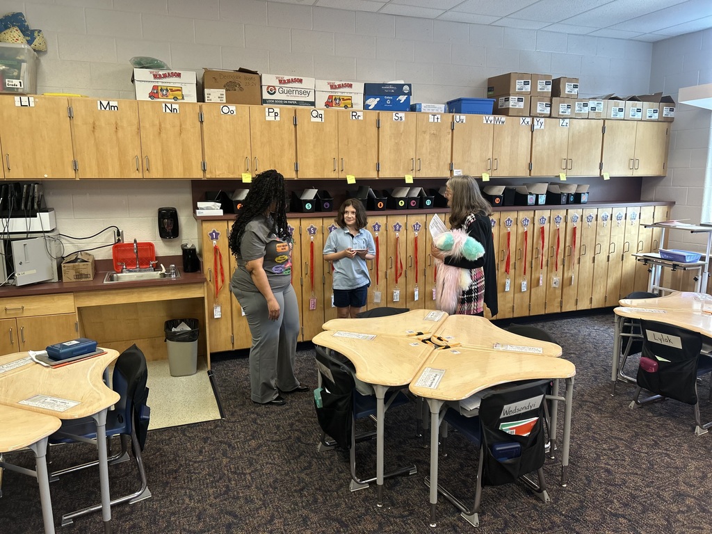 Two adults and one child are conversing in a conversation in a classroom setting with curved desks and wooden cabinets. 