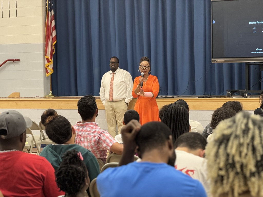 Ferguson PreK-8 Principal, Mrs. Lisa Love is speaking into a microphone in front of a stage, next to Ferguson PreK-8's Assistant Principal, Dr. Maurice Jones. An audience can be seen listening attentively, with a large screen in front of them.