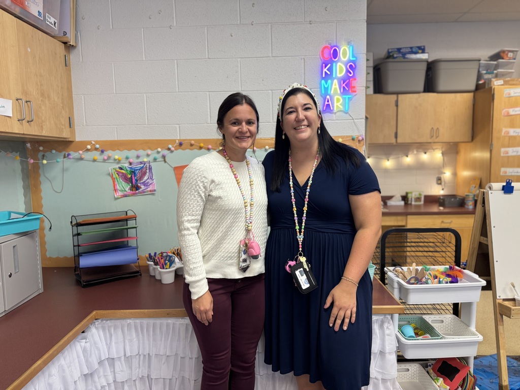 Two district staff members are posing together in a classroom setting. There are craft supplies and decor in the background, including a neon sign that reads "Cool Kids Make Art."