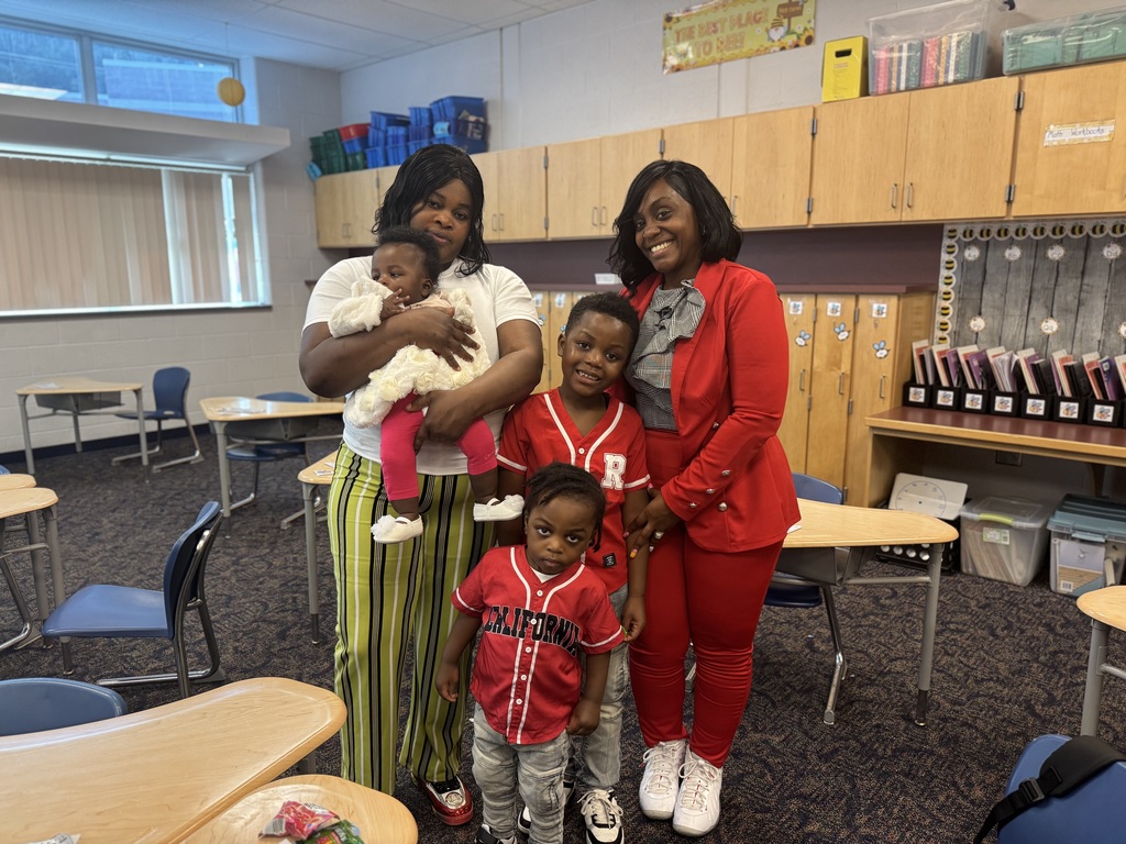 A group of five people, including two women, two young boys, and a baby pose happily in a classroom. The room has wooden cabinets and desks.