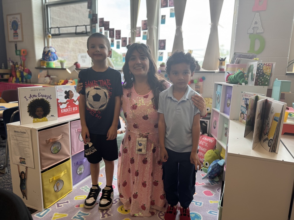 A district staff member is kneeling in between two young children in a classroom setting. Books and storage cubes can be seen in the background.