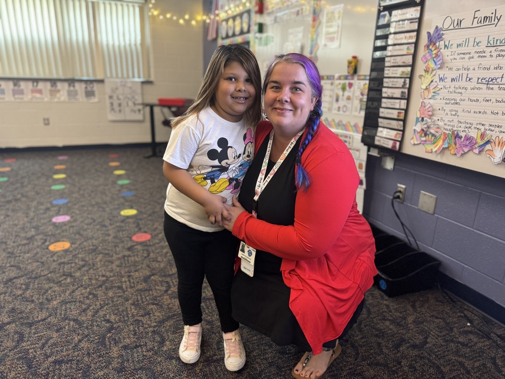 A district staff member is kneeling next to a student who is smiling in a classroom setting.