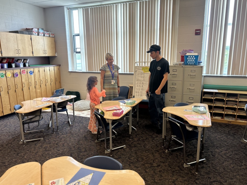 A district staff member, young child, and an adult are conversing in a classroom with wooden shelves, curved desks, and educational posters. 