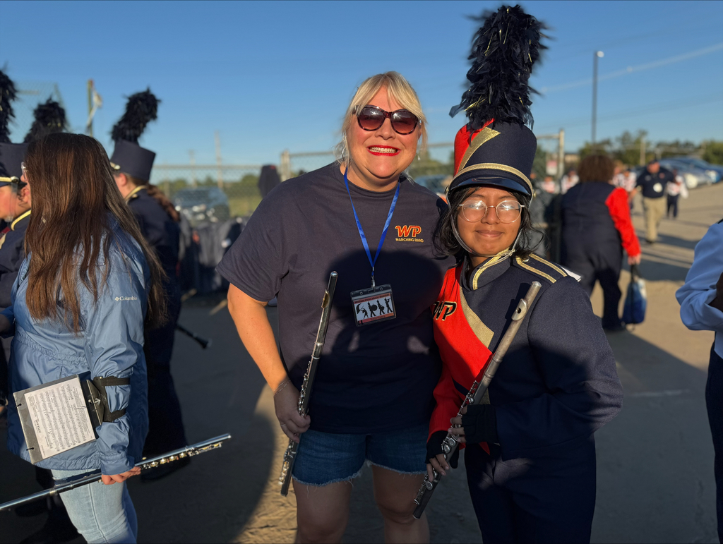 A district staff member and a York High band member posing together in a sunny outdoor setting, wearing uniforms and holding instruments, with other people in the background.